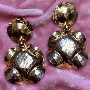 Peut inclure: Une paire de boucles d'oreilles dorées à la texture martelée. Les boucles d'oreilles sont composées de deux pièces, un disque rond et une forme plus irrégulière.
