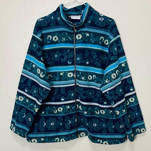 Może przedstawiać: Turkusowa i granatowa polarowa kurtka z kwiatowym i pasiastym wzorem. Kurtka ma pełny zamek błyskawiczny i wysoki kołnierz. Kwiatowy wzór zawiera białe i jasnoniebieskie akcenty. Kurtka wisi na białym wieszaku.