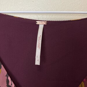 May include: A burgundy and gold floral print dress with a V-neckline. The dress has a Free People tag attached to the back of the neck. The tag reads "Made in Vietnam, Fabric: Made in Vietnam".