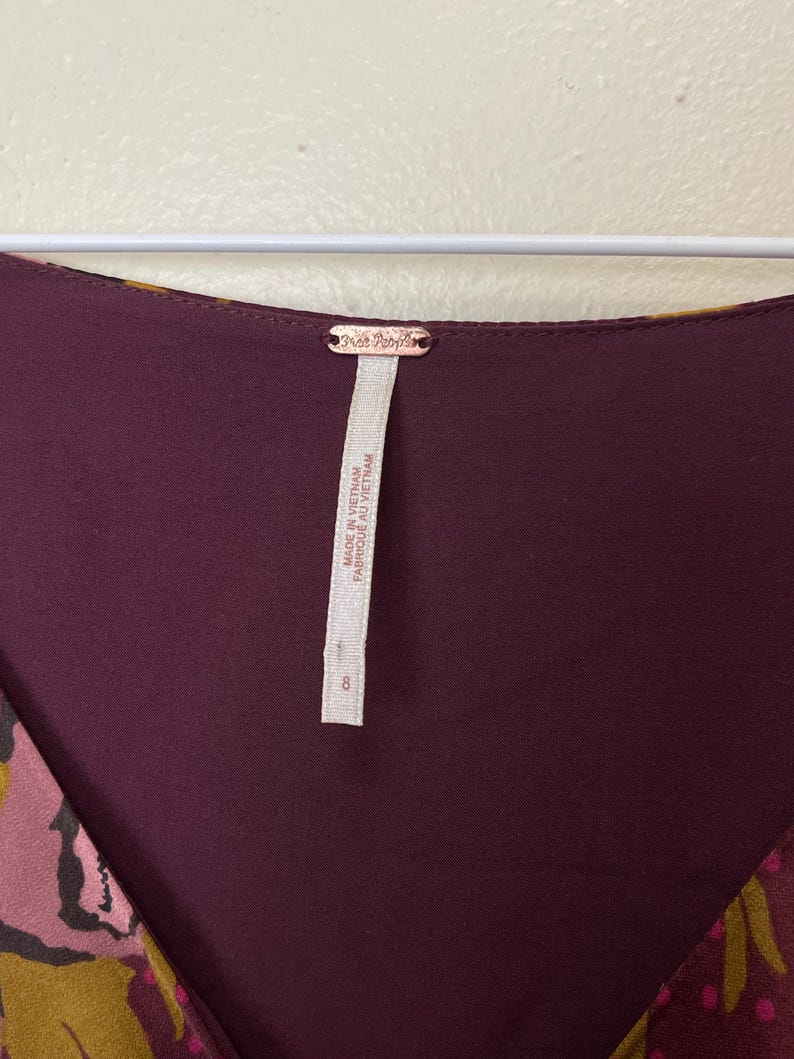 May include: A burgundy and gold floral print dress with a V-neckline. The dress has a Free People tag attached to the back of the neck. The tag reads "Made in Vietnam, Fabric: Made in Vietnam".