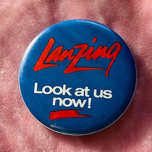 Puede incluir: Un botón azul y rojo con el texto "Lanzing Look at us now!" en letras blancas.