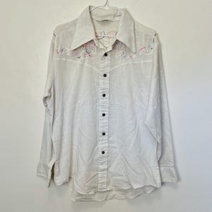 May include: Off-white long-sleeve shirt with a wide collar and black buttons. The shirt features embroidered swirls in red, pink, and blue on the collar. The shirt is hanging on a white hanger.