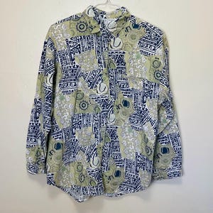 May include: A button-down shirt with a unique pattern. The shirt features a collage of geometric shapes and abstract designs in shades of olive green, navy blue, and white. The long-sleeved shirt is hanging on a white hanger against a white background.