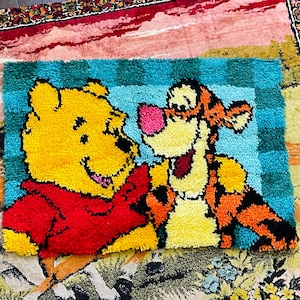 Könnte beinhalten: Ein rechteckiger, handgetufteter Teppich mit Winnie Puuh und Tigger. Der Teppich hat einen türkis-blauen Karomuster-Rand. Winnie Puuh ist gelb und trägt ein rotes Hemd. Tigger ist orange mit schwarzen Streifen.