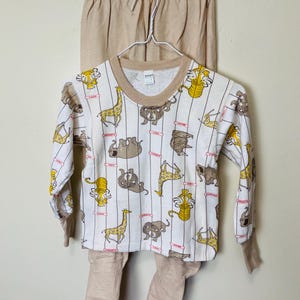 May include: A two-piece children's pajama set. The long-sleeved top is white with vertical stripes and cartoon animal illustrations in yellow and brown. The pants and the trim on the top are tan.