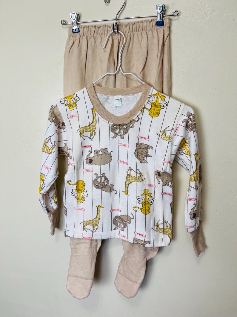 May include: A two-piece children's pajama set. The long-sleeved top is white with vertical stripes and cartoon animal illustrations in yellow and brown. The pants and the trim on the top are tan.