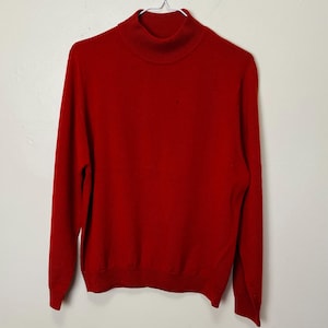 Puede incluir: Un jersey rojo liso de cuello alto colgado de una percha metálica. El jersey tiene mangas largas y un cuello acanalado. El material parece ser un tejido de punto suave. El jersey es de un tono rojo vibrante.