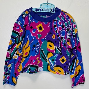Könnte beinhalten: Ein Vintage-Langarmshirt mit einem lebendigen abstrakten Muster in Blau, Lila, Gelb, Türkis und Pink. Das Shirt hat einen blauen Kragen und eine kleine Tasche. Zwei Knöpfe befinden sich auf der Schulter.