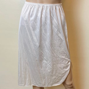 May include: A light peach-colored slip skirt with an elastic waistband. The skirt is made of a silky, flowing fabric and features a slit on the side. The skirt is knee-length.