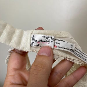 Pu&ograve; includere: Primo piano di un capo intimo color crema con dettagli in pizzo. L'etichetta recita "intimately free people" con accenti floreali. Il capo &egrave; tenuto da una mano.