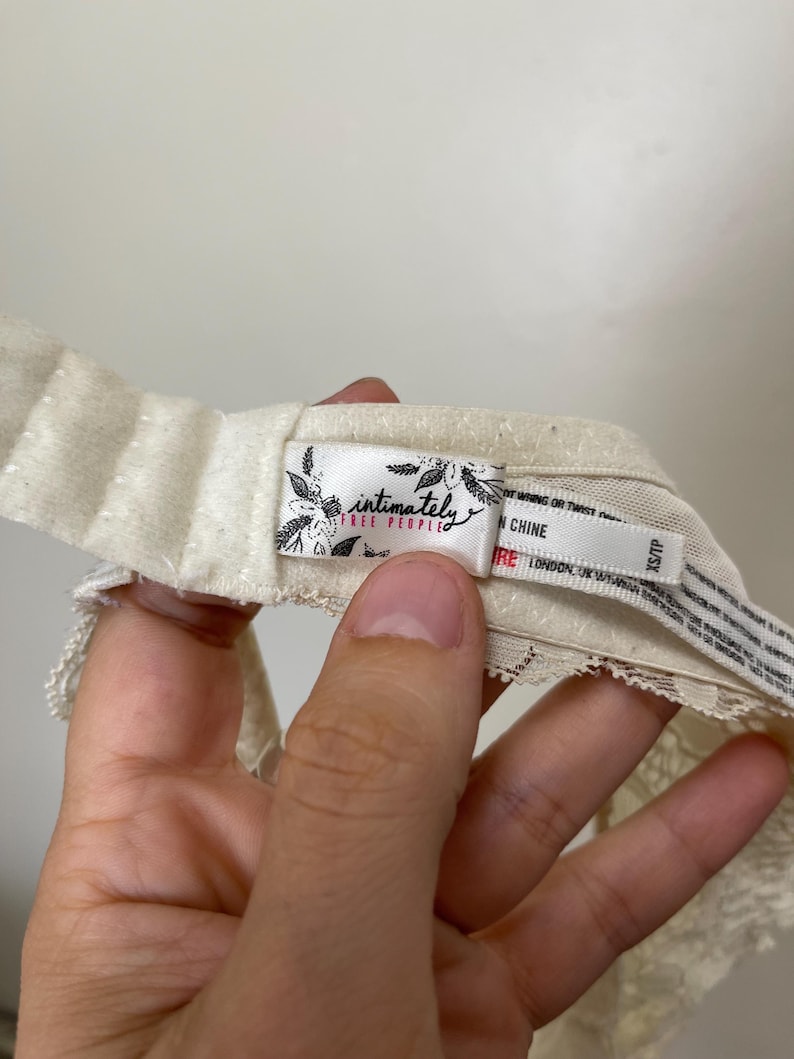 Pu&ograve; includere: Primo piano di un capo intimo color crema con dettagli in pizzo. L'etichetta recita "intimately free people" con accenti floreali. Il capo &egrave; tenuto da una mano.