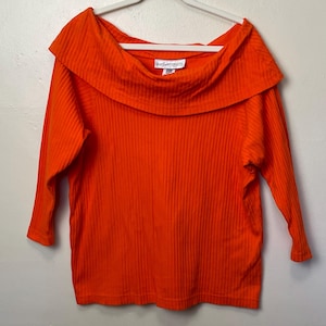 May include: Bright orange ribbed knit top with a wide, off-the-shoulder neckline and three-quarter sleeves. The top is hanging on a white hanger. The label reads "Yves Saint Objects".
