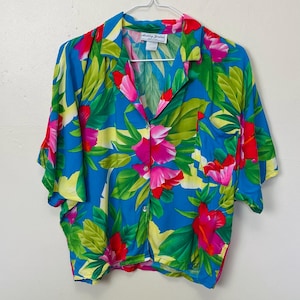May include: A short-sleeved button-down shirt with a vibrant floral pattern. The shirt features a bright blue background with large pink, red, and yellow flowers and green leaves. The label reads "Ashley Jordan".