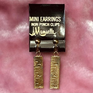 Puede incluir: Mini pendientes de oro con diseño de regla. Los pendientes son de clip y tienen el texto "MINI EARRINGS NON PINCH CLIPS Mamselle" en la tarjeta.