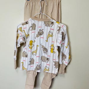 May include: A beige and white animal-themed pajama set. The long-sleeved top features a pattern of various animals, including giraffes and elephants, on a white background with vertical stripes. The set includes matching beige pants and footed bottoms.