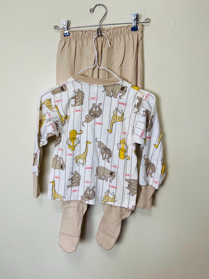 May include: A beige and white animal-themed pajama set. The long-sleeved top features a pattern of various animals, including giraffes and elephants, on a white background with vertical stripes. The set includes matching beige pants and footed bottoms.