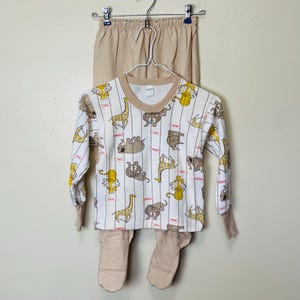 May include: A two-piece pajama set. The top is white with a repeating pattern of various animals in brown and yellow, with vertical stripes. The long-sleeved top has a brown neckline and cuffs. The pants are a solid tan color.