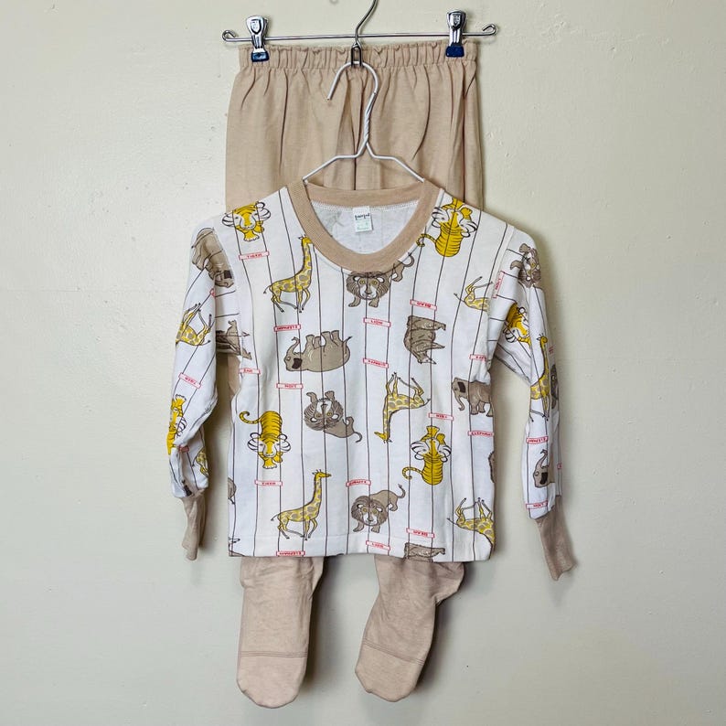 May include: A two-piece pajama set. The top is white with a repeating pattern of various animals in brown and yellow, with vertical stripes. The long-sleeved top has a brown neckline and cuffs. The pants are a solid tan color.