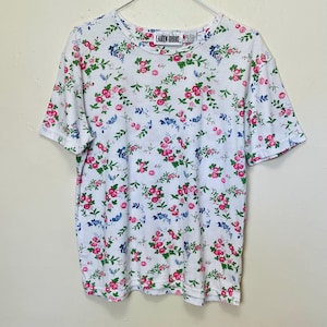 May include: White t-shirt with a floral pattern featuring pink roses, blue flowers, and green leaves. The shirt has a crew neck and short sleeves. The brand name "LAUREN BROOKE" is visible on the tag.