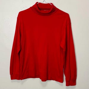 Puede incluir: Una camiseta roja de manga larga con cuello alto. La camisa tiene un cuello alto clásico y mangas largas con puños acanalados. La prenda está hecha de un material suave y está colgada de una percha blanca.