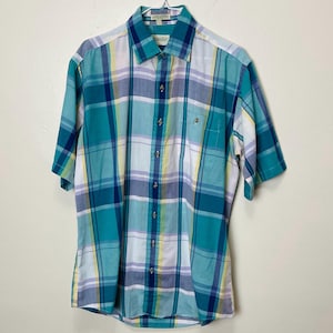 May include: A short-sleeved, multi-coloured plaid shirt in teal, blue, white, and yellow. The shirt features a button-down front, a chest pocket, and a collar. The shirt is displayed on a hanger against a white backdrop.
