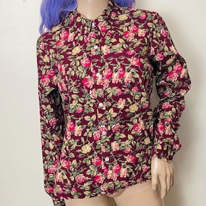 Puede incluir: Camisa abotonada de manga larga con fondo burdeos y estampado floral de rosas rosas y amarillas con hojas verdes. La camisa tiene cuello clásico y botones blancos.