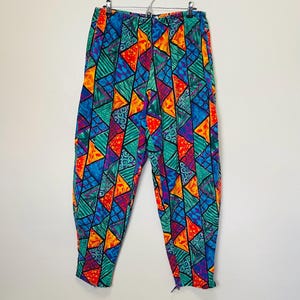 May include: Vibrant geometric patterned trousers in a mix of shapes and designs, featuring blue, green, orange, red, and purple hues. The trousers are suspended on a silver hanger against a white backdrop.
