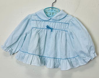 May include: A light blue baby blouse with white polka dots and a ruffled hem. The blouse has a Peter Pan collar and a bow at the neckline.
