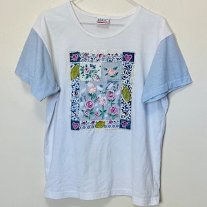 以下が含まれることがあります： 白地に青と白のストライプの袖が付いたTシャツ。Tシャツの前身頃には、パッチワークのデザインの花柄がプリントされています。プリントには、ピンク、ブルー、グリーン、ホワイトの花が含まれています。タグには「J-BASIC」の文字がプリントされています。