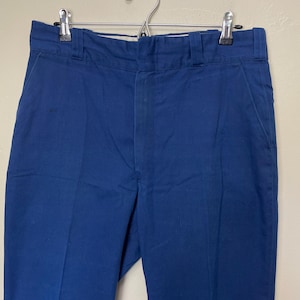 Puede incluir: Un pantalón azul marino colgado de una percha metálica. El pantalón tiene un diseño clásico de pierna recta con bolsillos laterales y cierre de botón y cremallera. La tela parece ser un algodón o mezcla resistente.