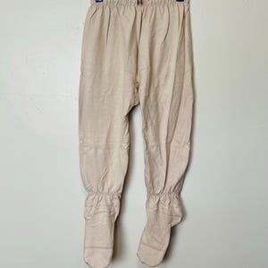 May include: Beige-colored footed pants hanging on a metal hanger. The pants have a gathered waist and elasticized ankles. The fabric appears to be a soft, knit material. The pants are a neutral color.
