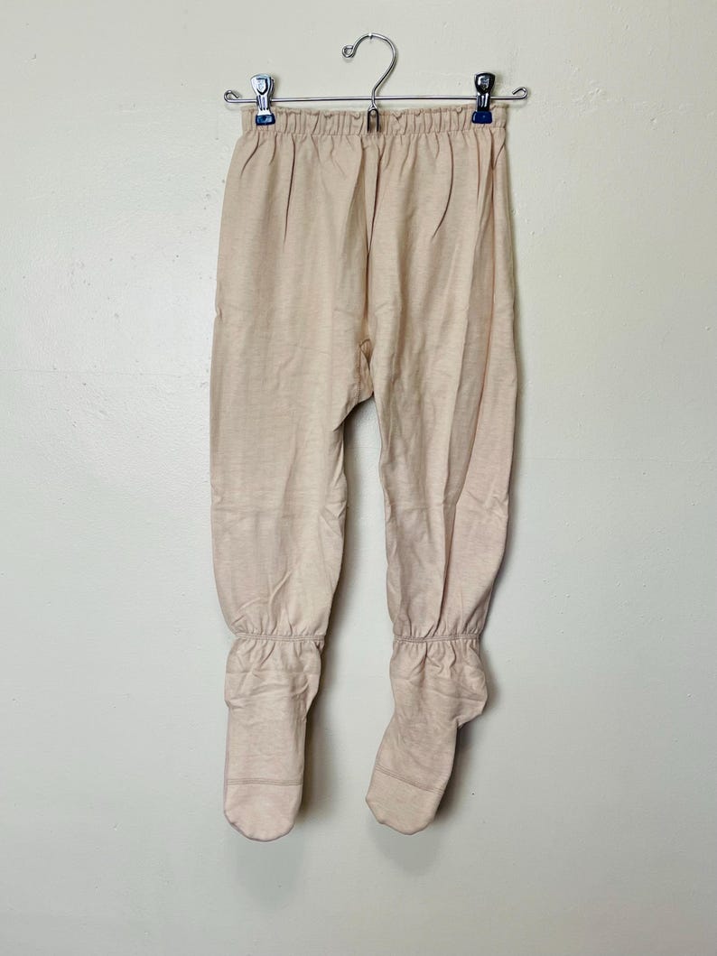 May include: Beige-colored footed pants hanging on a metal hanger. The pants have a gathered waist and elasticized ankles. The fabric appears to be a soft, knit material. The pants are a neutral color.