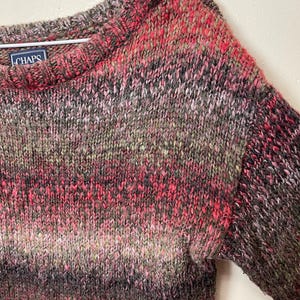 May include: A multi-colored knit sweater with a crew neck. The sweater features a blend of pink, red, black, and gray yarns. The brand label "CHAPS" is visible on the upper left side. The sweater is hanging on a metal hanger.