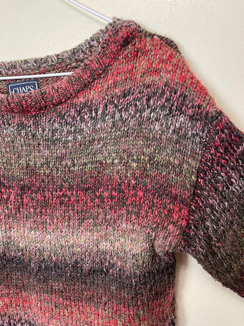 May include: A multi-colored knit sweater with a crew neck. The sweater features a blend of pink, red, black, and gray yarns. The brand label "CHAPS" is visible on the upper left side. The sweater is hanging on a metal hanger.