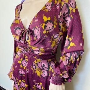 May include: A long-sleeved dress with a floral print in shades of pink, yellow, and brown on a burgundy background. The dress has a V-neckline with a tie detail at the front and a fitted bodice. The sleeves are long and have a gathered elastic band at the cuffs.