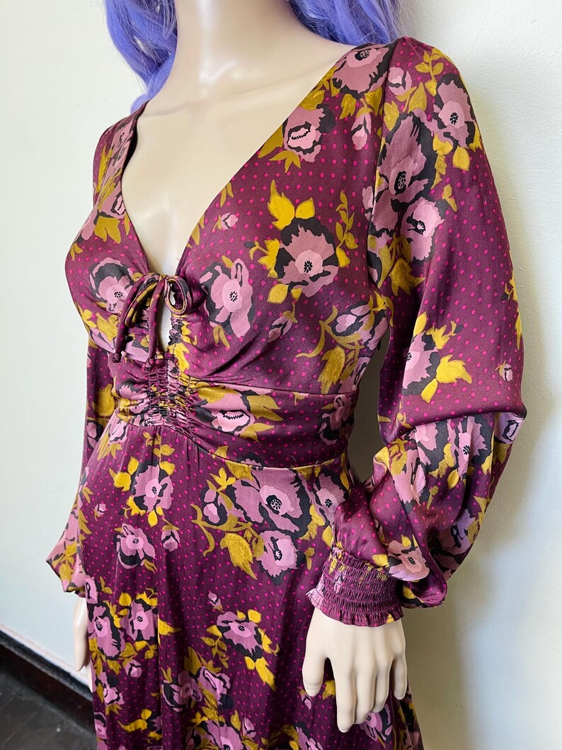 May include: A long-sleeved dress with a floral print in shades of pink, yellow, and brown on a burgundy background. The dress has a V-neckline with a tie detail at the front and a fitted bodice. The sleeves are long and have a gathered elastic band at the cuffs.