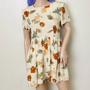 May include: A white dress with a floral print featuring brown flowers, green leaves, and gold geometric shapes. The dress has a pleated skirt and short sleeves.