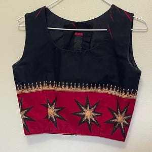 May include: Sleeveless black top with a square neckline. The bottom portion features a red band with large starburst designs in black and gold. The top is hanging on a white hanger.