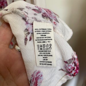 Op de afbeelding: Close-up van een witte stof met een bloemenpatroon in roze en rode tinten. Een label met wasvoorschriften is zichtbaar. De stof is gemaakt van 100% rayon.