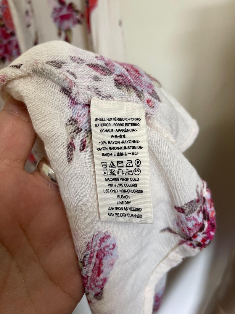 Op de afbeelding: Close-up van een witte stof met een bloemenpatroon in roze en rode tinten. Een label met wasvoorschriften is zichtbaar. De stof is gemaakt van 100% rayon.