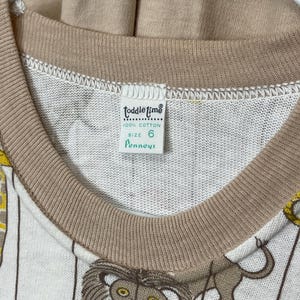 May include: Vintage children's long-sleeve shirt with a circus lion print. The shirt is white with vertical stripes and features a brown collar and cuffs. The tag reads "Toddle Time" and "100% Cotton", size 6.