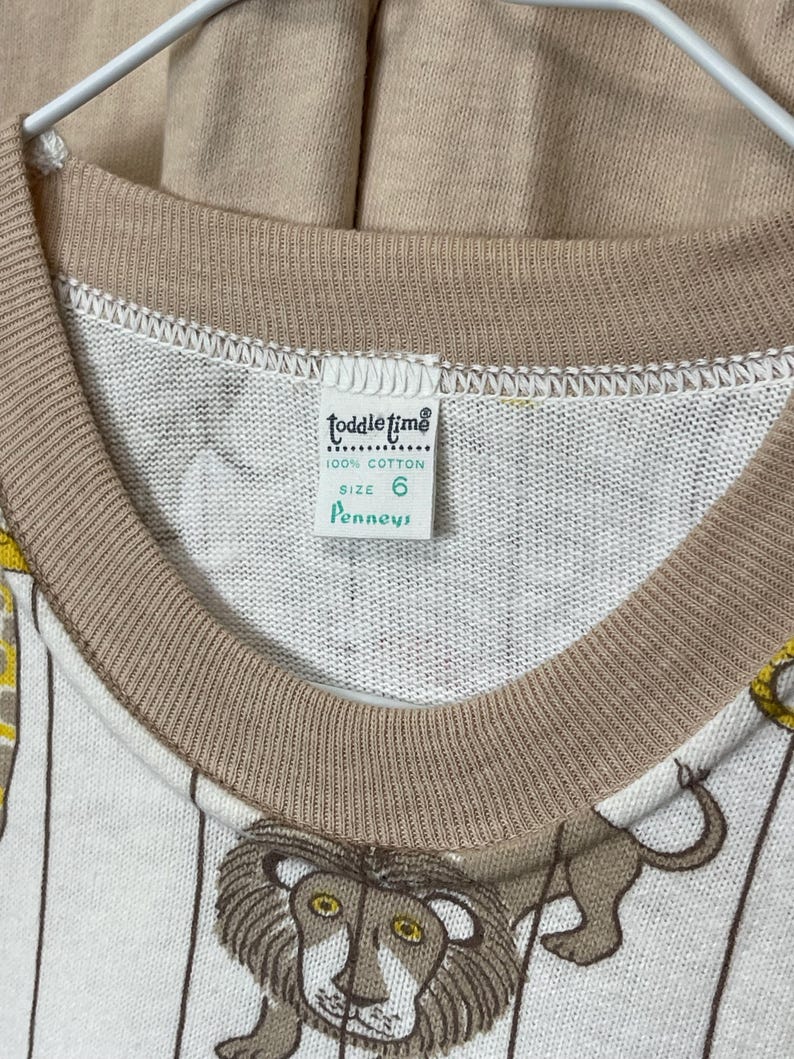 May include: Vintage children's long-sleeve shirt with a circus lion print. The shirt is white with vertical stripes and features a brown collar and cuffs. The tag reads "Toddle Time" and "100% Cotton", size 6.