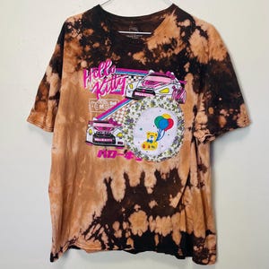 May include: A tie-dye t-shirt with a Hello Kitty graphic. The graphic features two race cars, a rainbow, and a teddy bear holding balloons. The shirt has a brown and black tie-dye pattern.