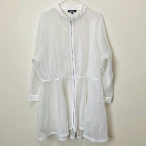 Puede incluir: Chaqueta de malla blanca con cuello alzado y cremallera completa. La chaqueta tiene mangas largas y un bajo acampanado en capas, similar a una falda. La tela parece ligera y semitransparente, adecuada para superponer. La chaqueta está colgada en una percha.
