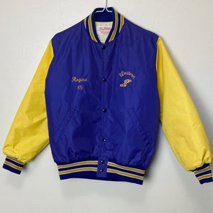 Op de afbeelding: Een blauw en geel varsity jack met de tekst "Regina 85" en "Western" geborduurd op de voorkant. Het jack heeft een goud en blauw gestreepte kraag en manchetten.