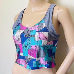 May include: A sleeveless crop top with a colorful geometric pattern in shades of blue, pink, gray, and white. The top has a scoop neckline and gray striped shoulder straps. The fabric appears to be a lightweight material.
