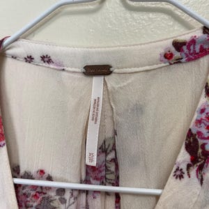 Op de afbeelding: Cr&egrave;mekleurige blouse met een bloemenpatroon in roze en bordeaux tinten. De blouse heeft een V-hals en hangt aan een witte hanger. Het label luidt "Free People" en "Made in Vietnam".
