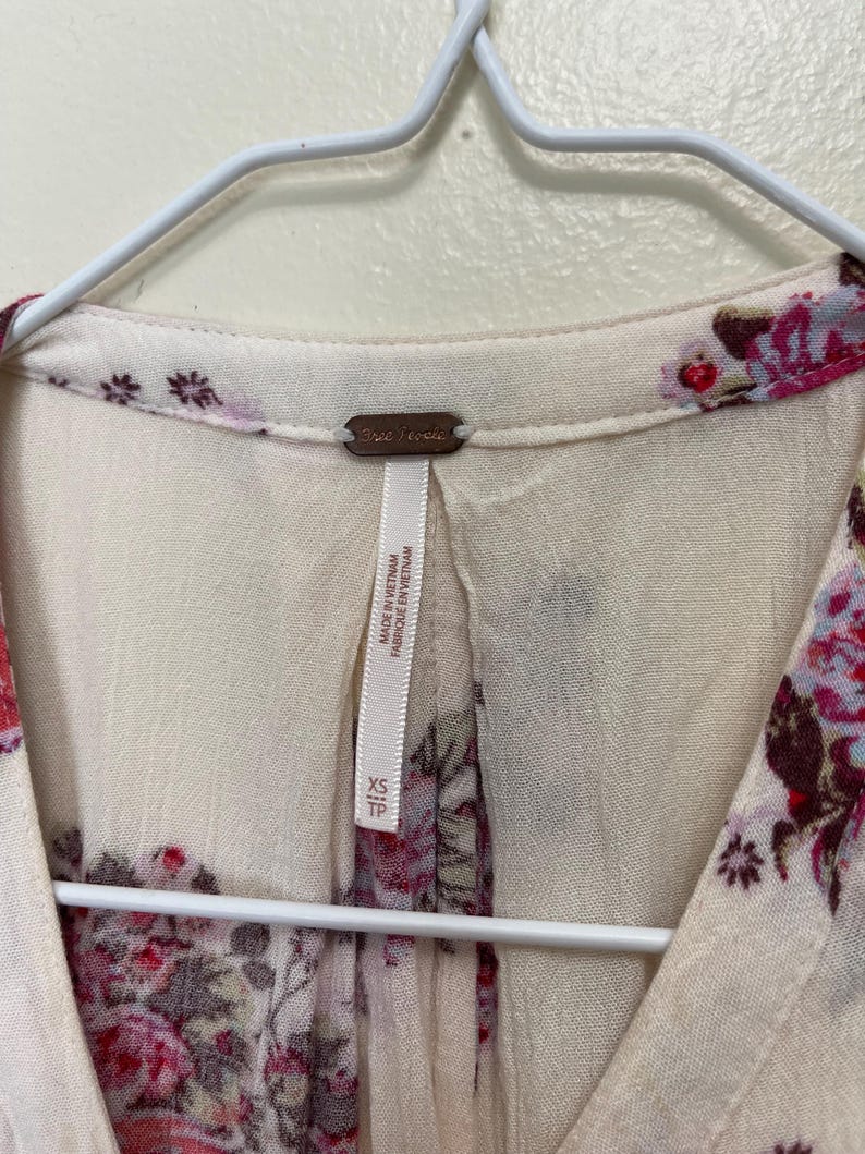 Op de afbeelding: Cr&egrave;mekleurige blouse met een bloemenpatroon in roze en bordeaux tinten. De blouse heeft een V-hals en hangt aan een witte hanger. Het label luidt "Free People" en "Made in Vietnam".