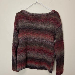 May include: A multi-colored sweater with a scoop neck, hanging on a white metal hanger. The sweater features horizontal stripes of red, brown, and gray. The knitwear appears soft and cozy.