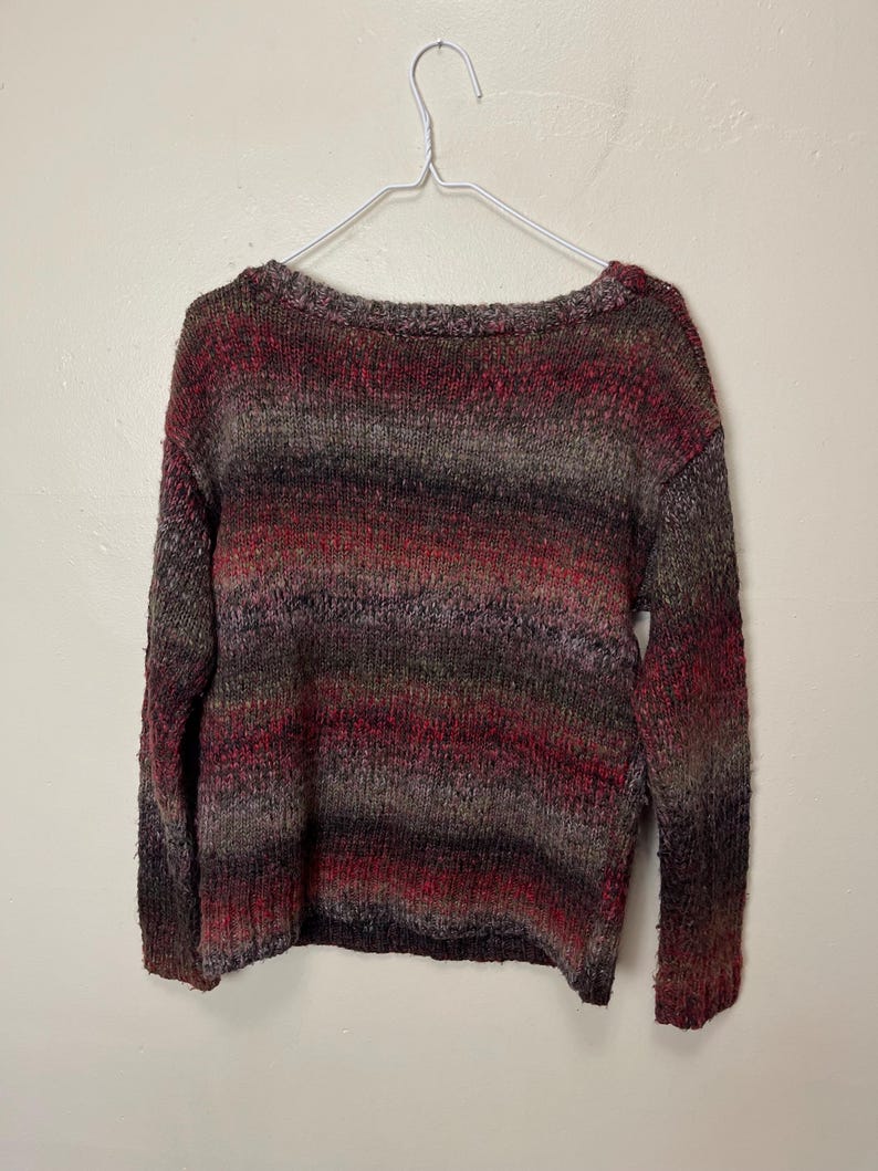 May include: A multi-colored sweater with a scoop neck, hanging on a white metal hanger. The sweater features horizontal stripes of red, brown, and gray. The knitwear appears soft and cozy.